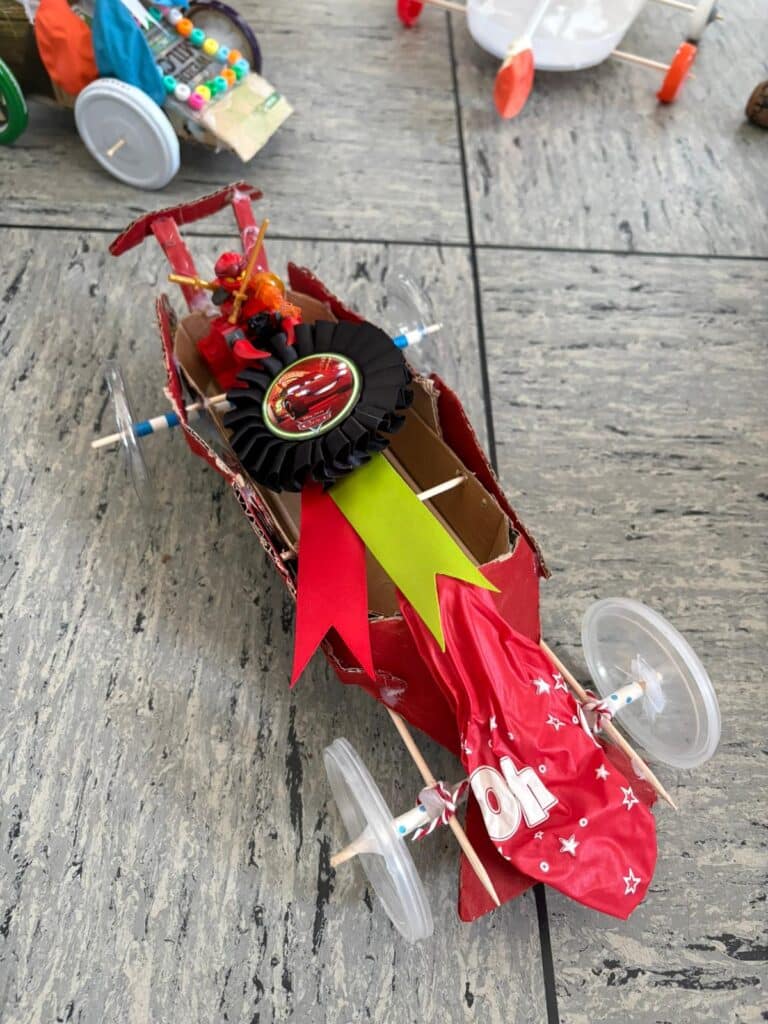Ein selbstgebauter Spielzeug-Rennwagen aus Pappe, Flaschenverschlüssen und Plastikrädern, verziert mit rotem Papier, Bändern und einer kleinen Fahrerfigur.