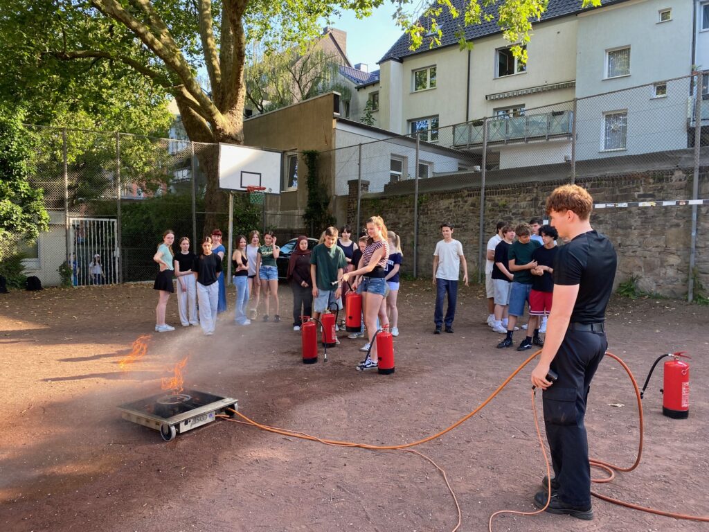 Eine Gruppe von Jugendlichen beobachtet, wie eine Person bei einer Brandschutzvorführung im Freien mit einem Feuerlöscher die Flammen auf einem Tablett löscht.
