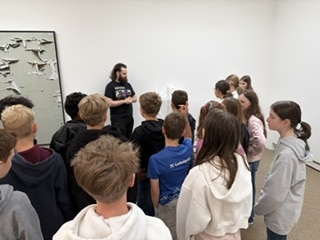Eine Gruppe von Kindern hört einem Mann zu, der in einer Kunstgalerie spricht, während hinter ihnen Kunstwerke an einer weißen Wand hängen.