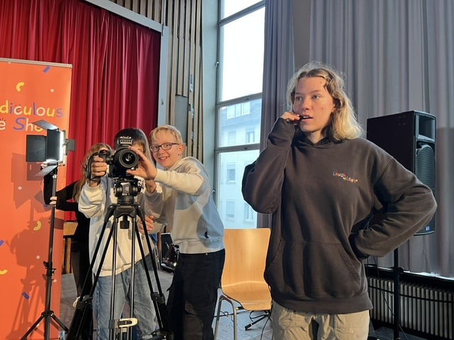 Drei junge Leute filmen mit einer Videokamera in einem Raum; zwei bedienen die Kamera, während einer davor steht und nachdenklich mit der Hand am Kinn dasitzt.