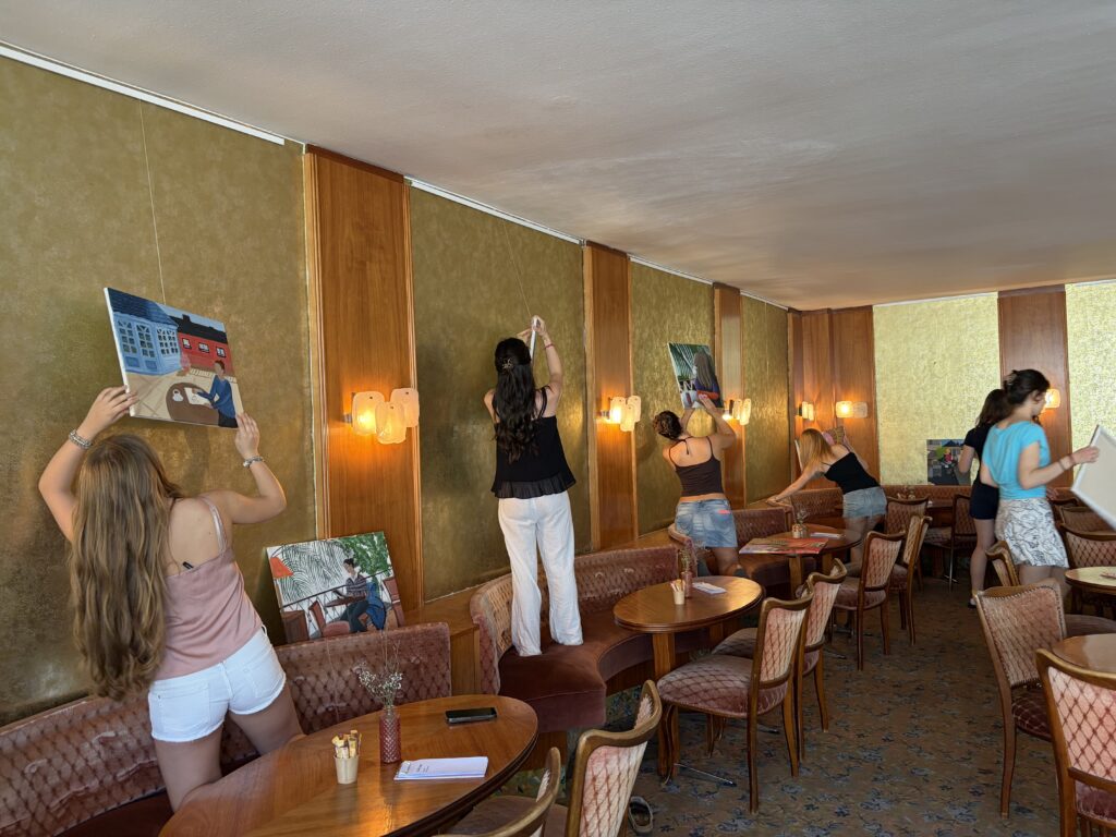 Sechs Frauen hängen Gemälde an eine Wand in einem Raum mit Holzvertäfelung, pfirsichfarbenen Stühlen und runden Tischen. Einige stellen sich auf Stühle, um auch die höheren Stellen zu erreichen.