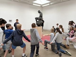 Eine Gruppe von Kindern imitiert eine sitzende Statue eines Denkers in der Mitte einer Kunstgalerie mit weißen Wänden und einem Oberlicht.