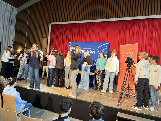 Eine Gruppe von Kindern auf der Bühne nimmt an einem Workshop teil, während andere filmen und Fotos machen; die sitzenden Zuschauer verfolgen das Geschehen vor einem roten Vorhang als Hintergrund.