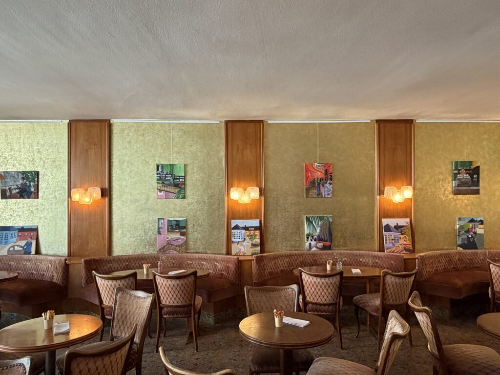 Ein gemütliches Café im Vintage-Stil mit Holzstühlen, runden Tischen, gepolsterten Sitznischen, Wandlampen und bunten, gerahmten Fotos an den grünlichen Wänden.