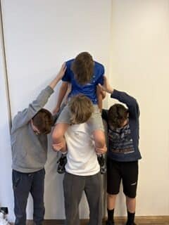 Vier Jungen posieren in einem Haus; ein Junge in Blau sitzt auf den Schultern eines anderen, während zwei andere auf jeder Seite stehen und den Jungen, der oben sitzt, berühren.