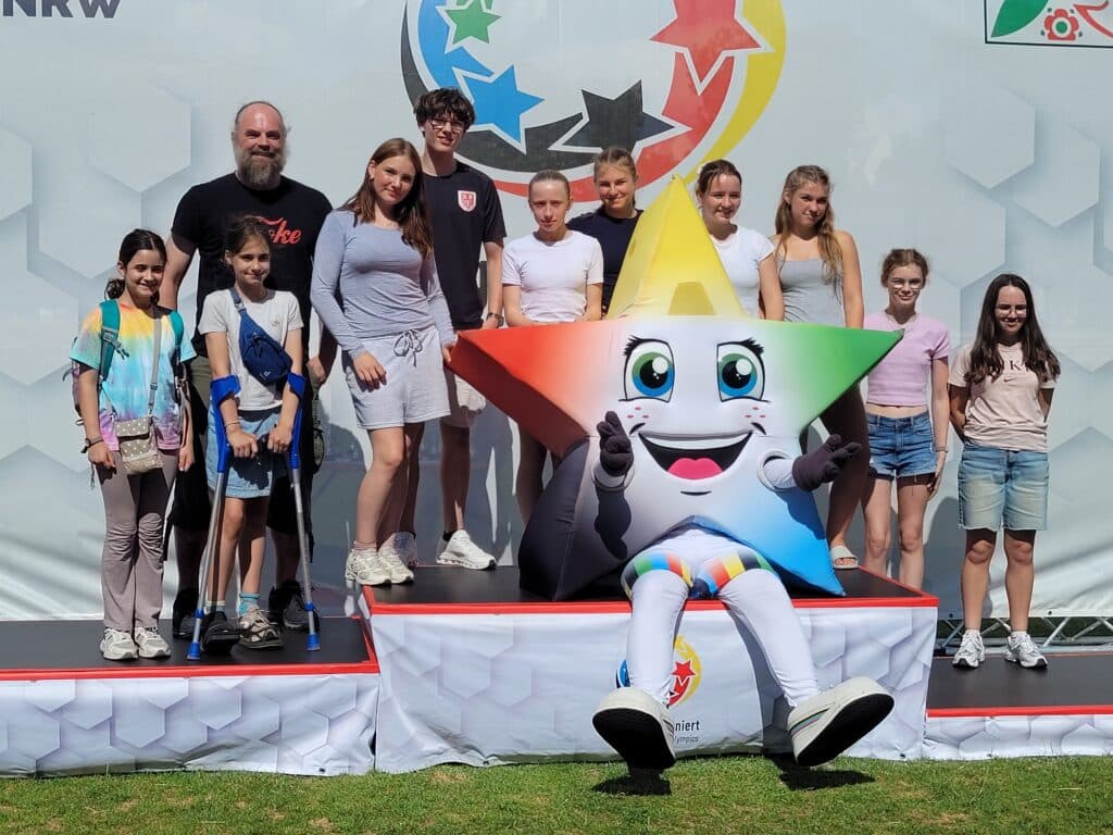 Eine Gruppe von Erwachsenen und Kindern steht und sitzt an einem sonnigen Tag auf einer Freiluftbühne um ein buntes Stern-Maskottchen herum und lächelt in die Kamera.