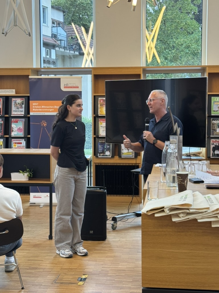 Ein Mann spricht in ein Mikrofon, während er neben einer jungen Frau in einer Bibliothek steht; im Hintergrund sind Bücher, Zeitschriften und ein Fernsehbildschirm zu sehen.