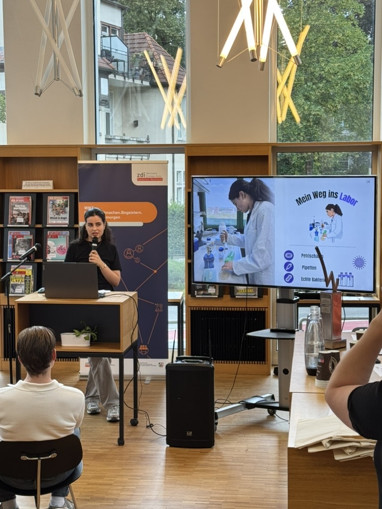 Eine Frau hält in einer Bibliothek einen Vortrag, neben ihr steht eine Diashow mit dem Titel „Mein Weg ins Labor“; die Zuhörer sitzen und hören zu.
