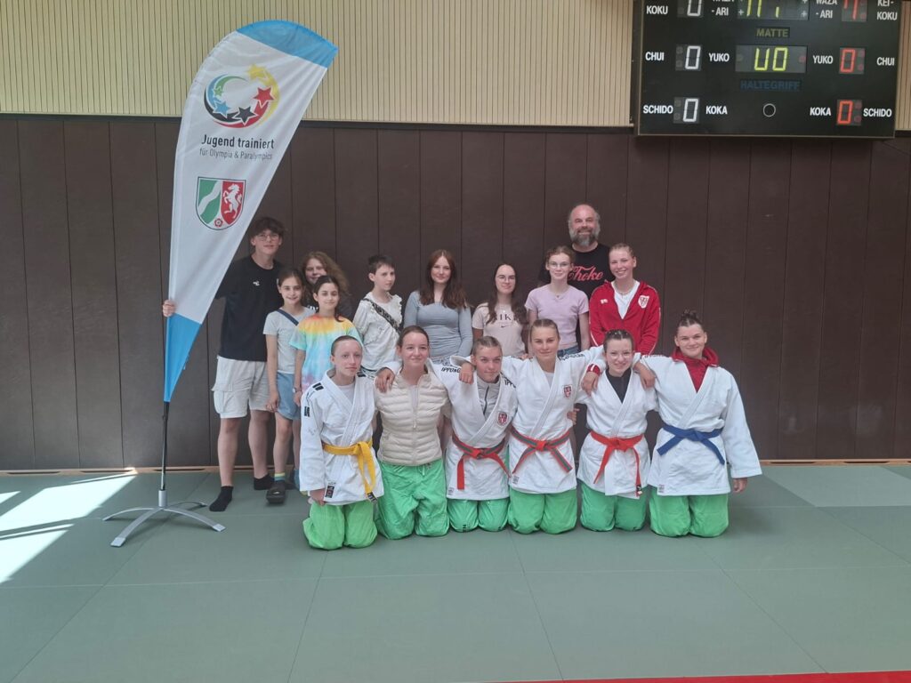 Eine Gruppe von Jugendlichen und Erwachsenen posiert in einem Raum auf einer Judomatte, einige tragen Judokleidung und farbige Gürtel, neben einer „Jugend trainiert“-Fahne.