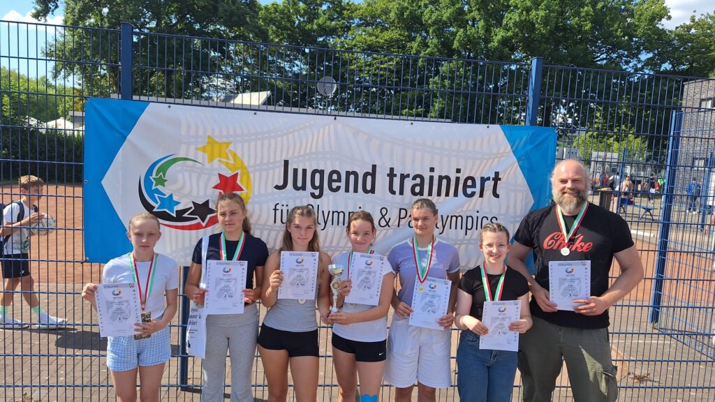 Sieben Personen – sechs Mädchen und ein Mann – stehen vor einem „Jugend trainiert“-Banner und halten jeweils Urkunden und Medaillen in den Händen; ein Mädchen hält einen Pokal in der Hand.