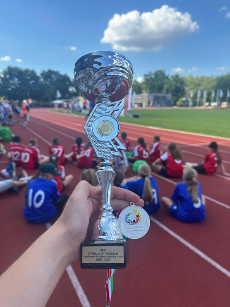 Auf einer Sportbahn hält eine Hand eine silberne Trophäe und eine Medaille in der Hand, im Hintergrund sitzen Kinder in bunten Sportuniformen.