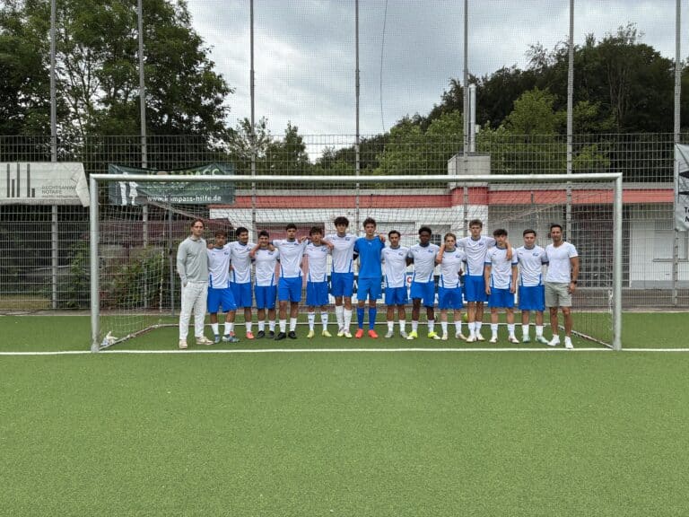 Eine Fußballmannschaft mit fünfzehn Spielern, die weiße Trikots und blaue Shorts tragen, steht in einer Reihe unter einem Torpfosten auf einem grünen Feld, links davon steht der Trainer.
