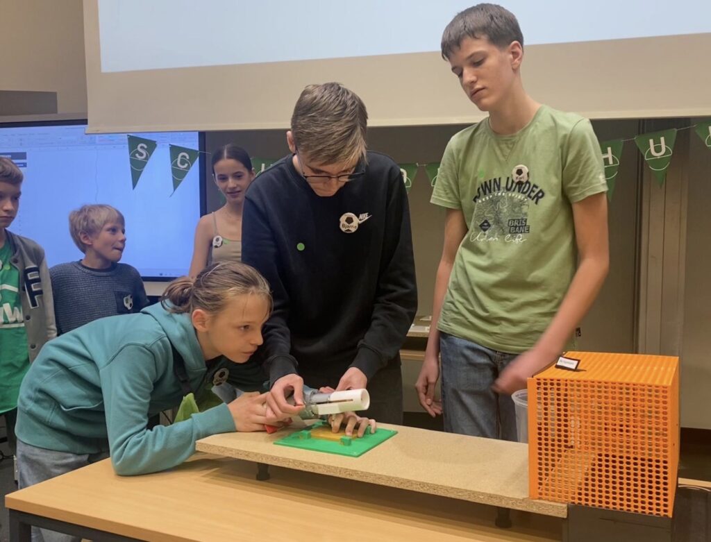 Drei Schüler konzentrieren sich auf ein kleines wissenschaftliches Experiment mit einer Spritze, während andere zuschauen. Sie befinden sich in einem Klassenzimmer mit einer Projektionswand und einem geschmückten Tisch.