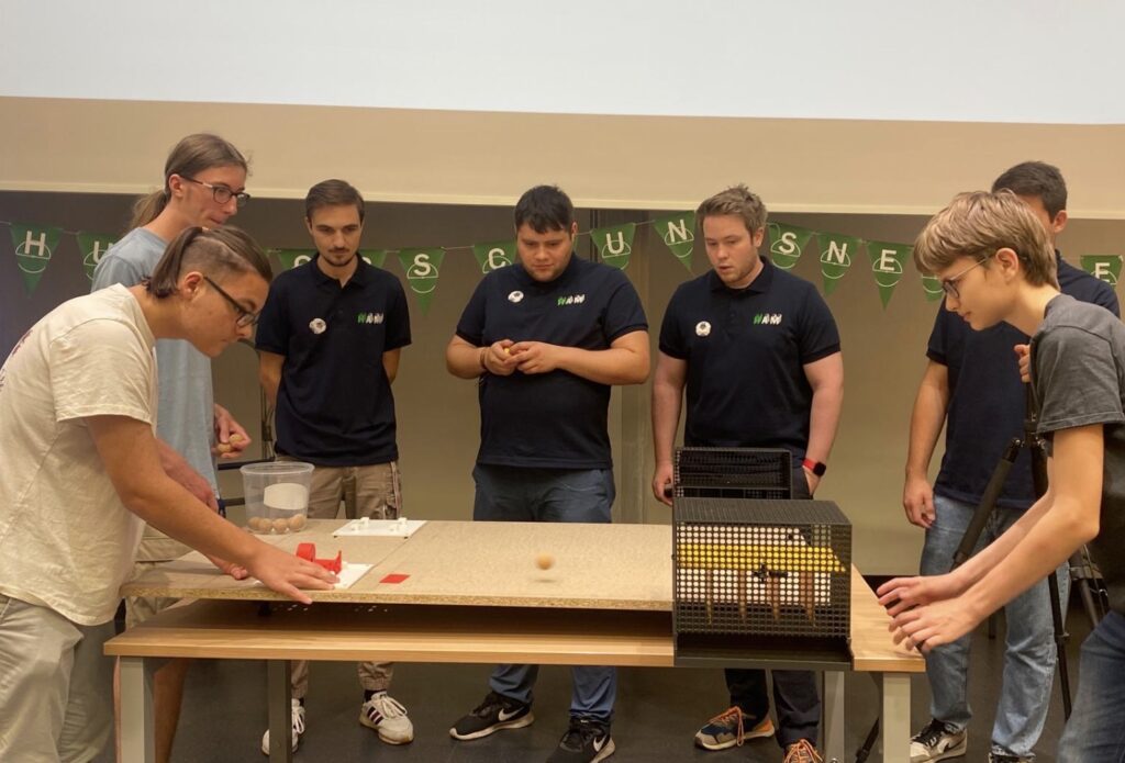 Sieben Jugendliche beobachten, wie eine Person einen Gegenstand auf einem Brett bewegt – möglicherweise im Rahmen einer Robotik- oder Technikvorführung in einem Klassenzimmer.