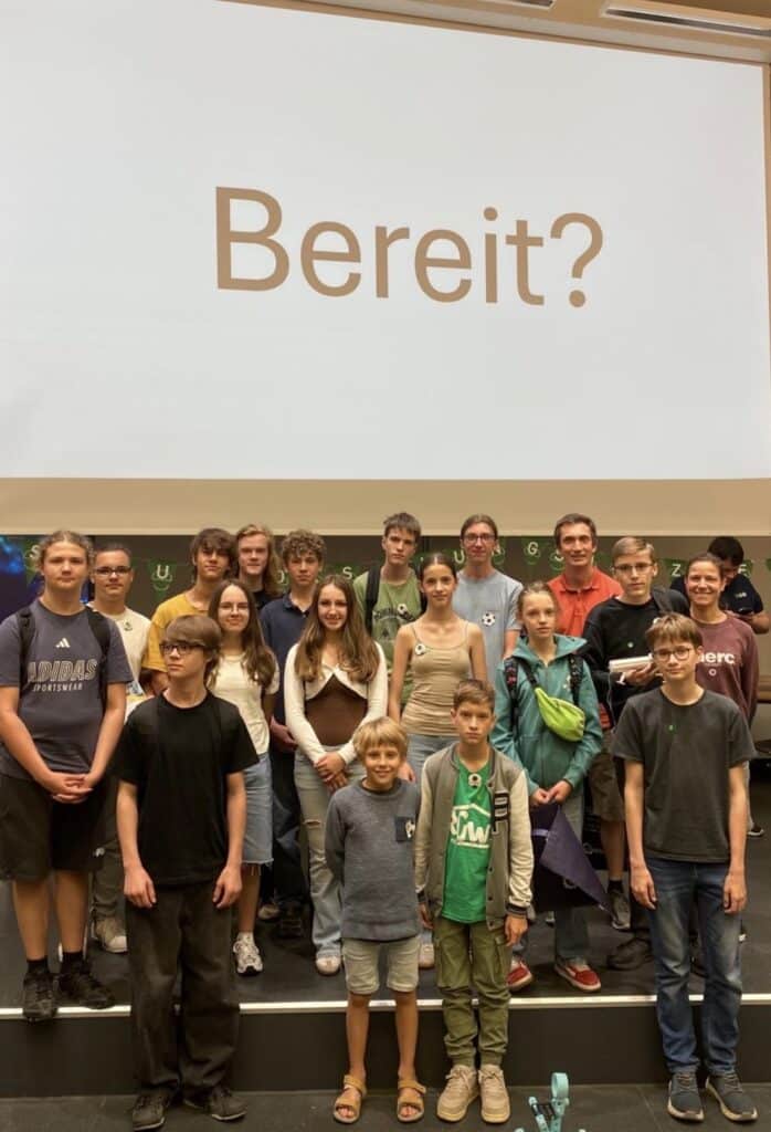 Eine Gruppe junger Menschen steht auf einer Bühne unter einer großen Leinwand, auf der das deutsche Wort „Bereit?“ zu sehen ist, und posiert lächelnd für die Kamera.