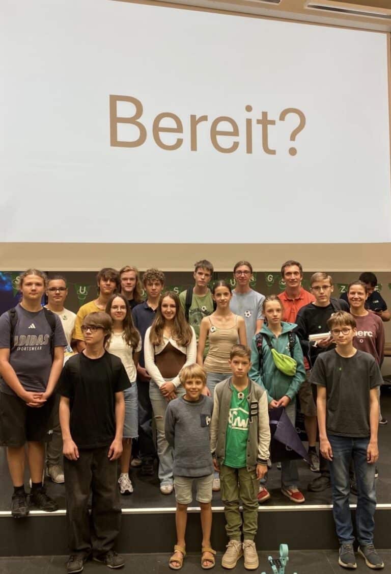 Eine Gruppe junger Menschen steht auf einer Bühne unter einer großen Leinwand, auf der das deutsche Wort „Bereit?“ zu sehen ist, und posiert lächelnd für die Kamera.