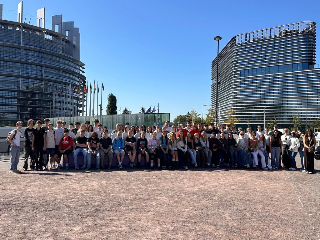 Eine große Gruppe von Menschen posiert an einem sonnigen Tag im Freien vor modernen Glasgebäuden und Fahnen.