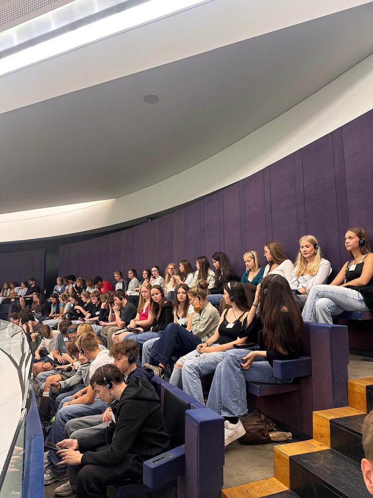 Eine Gruppe von Menschen, überwiegend junge Erwachsene, sitzt auf den gestuften Sitzreihen in einem modernen Auditorium; einige tragen Kopfhörer und hören aufmerksam zu.