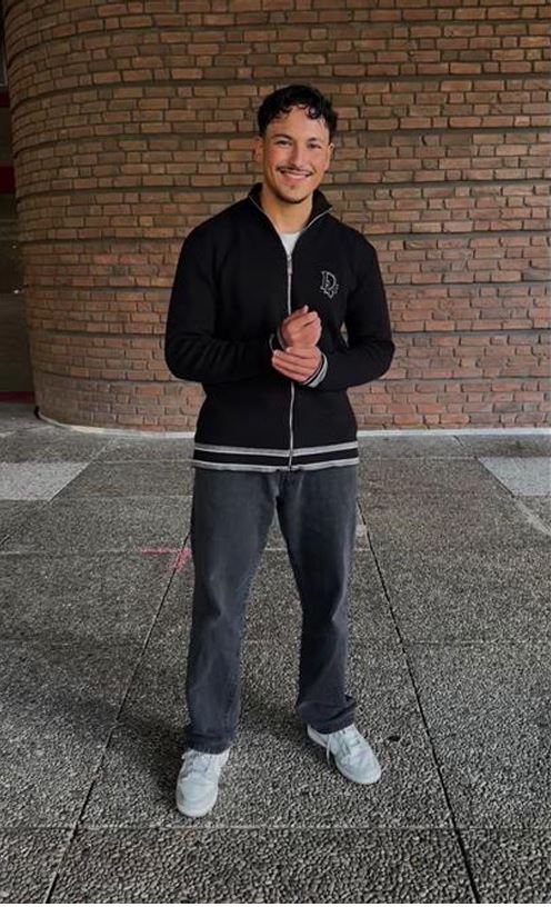 Ein Mann in einer schwarzen Jacke, grauen Jeans und weißen Sneakers steht lächelnd vor einer Backsteinmauer auf einem gepflasterten Bereich.