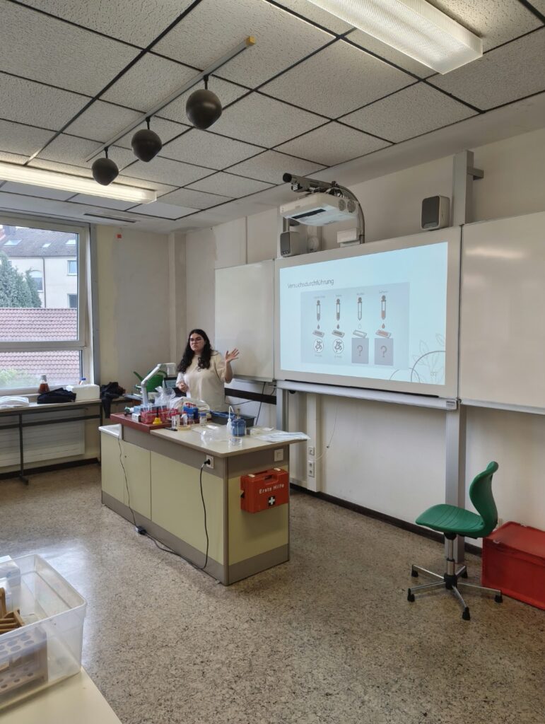 Eine Frau führt in einem Klassenzimmer ein wissenschaftliches Experiment vor; sie steht neben einem Labortisch mit Materialien und einer Leinwand, auf der Diagramme zu sehen sind.