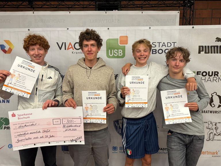 Vier junge Männer stehen in einer Reihe und halten ihre Marathon-Teilnahmebescheinigungen in den Händen; der zweite von links hält außerdem einen großen Werbescheck über 300 Euro in der Hand.