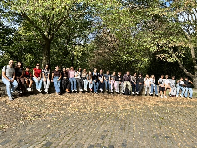 Eine große Gruppe von Schülern und Erwachsenen sitzt in einer langen Reihe auf einer Steinmauer in einem Park.